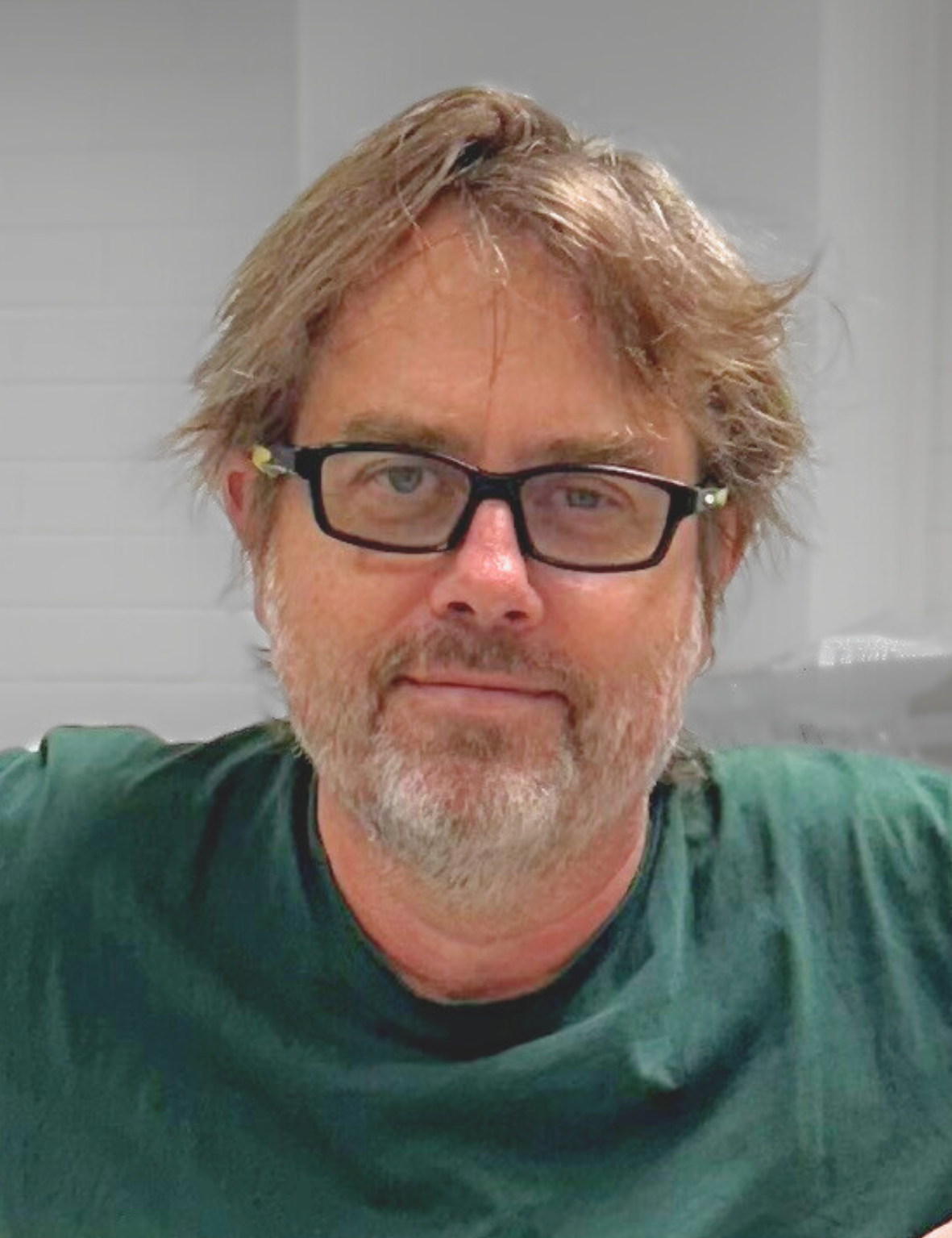 Man in a green shirt with black glasses. He has a grey beard and is smiling at the camera