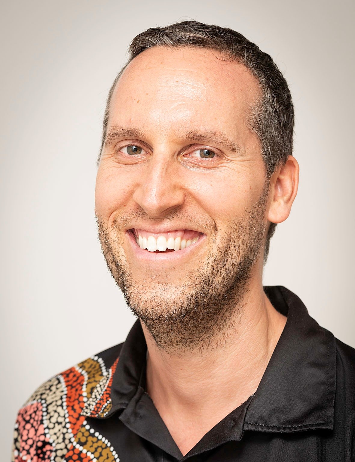 Portrait photo of a man wearing a black collared shirt with an Indigenous print on the shoulder. He is smiling at the camera. 