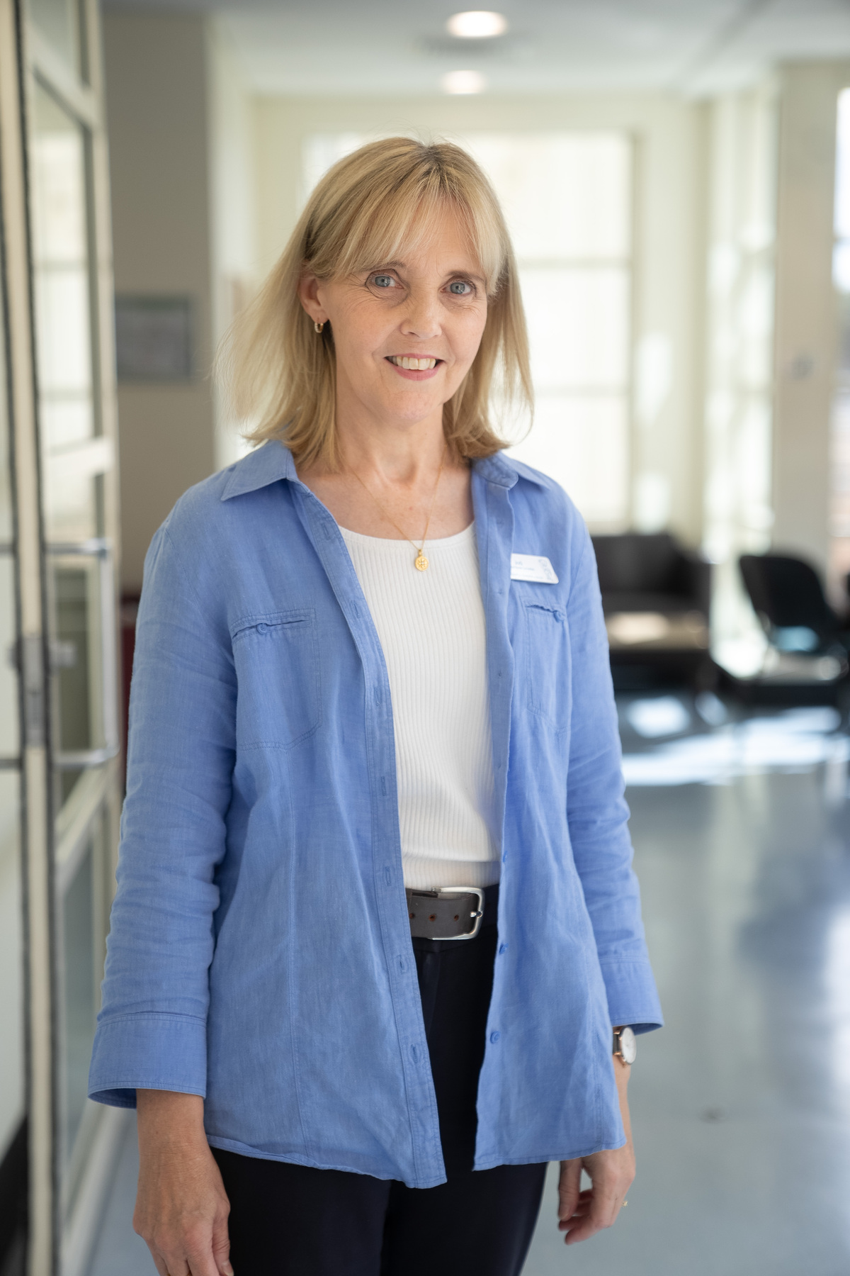 Woman in a white shirt and blue blazer is standing in a bright hospital setting. She has a blonde bob and is smiling at the camera. 