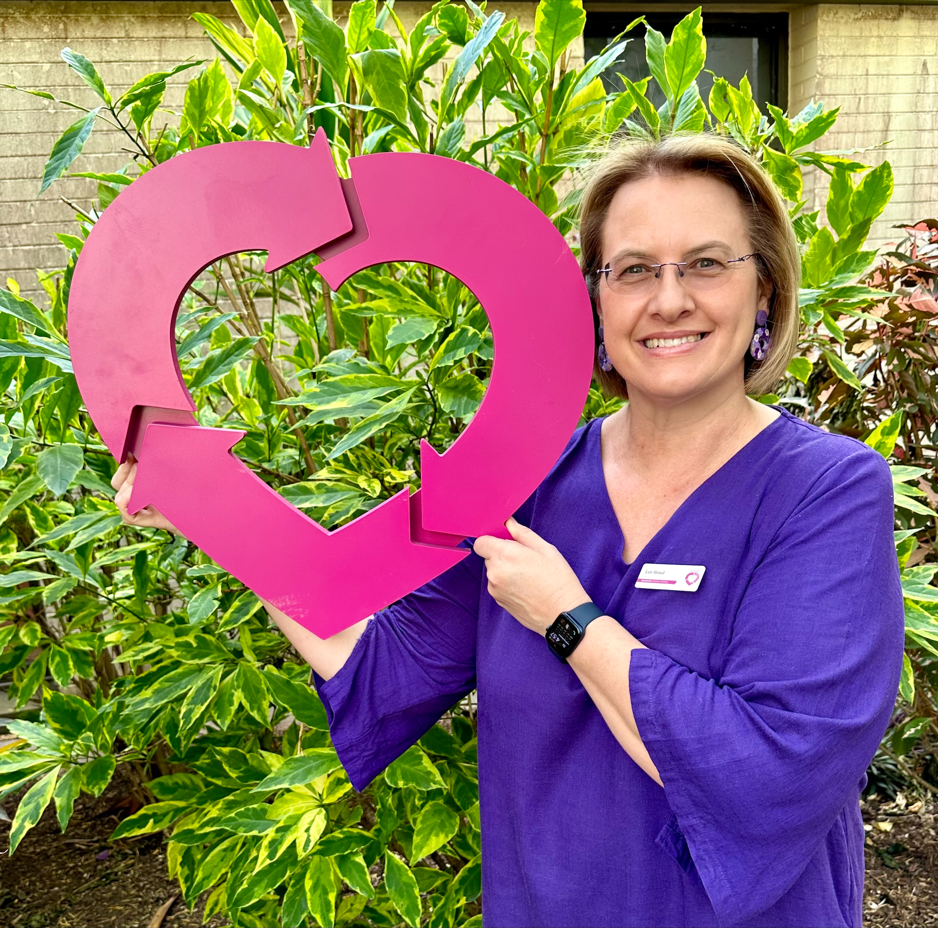 Woman in a purple shirt holding a large DonateLife heart. She is standing in front of a green bush and is smiling at the camera