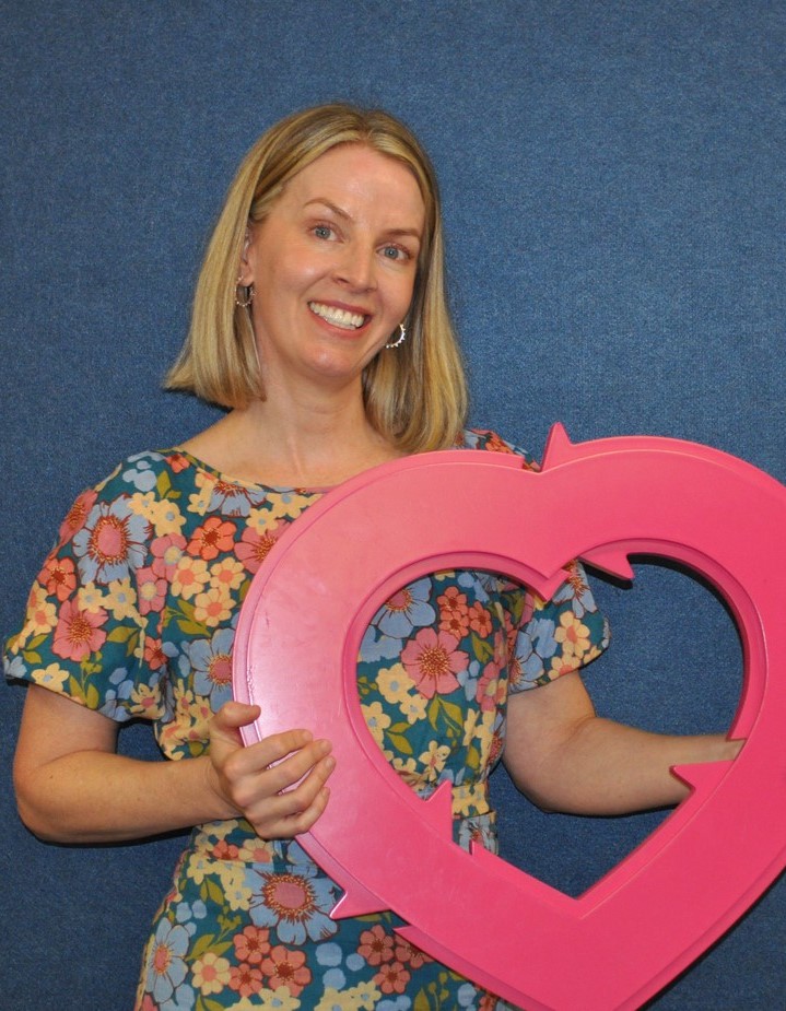 Woman with a blonde bob is wearing a patterned dress and is holding a pink DonateLife heart.