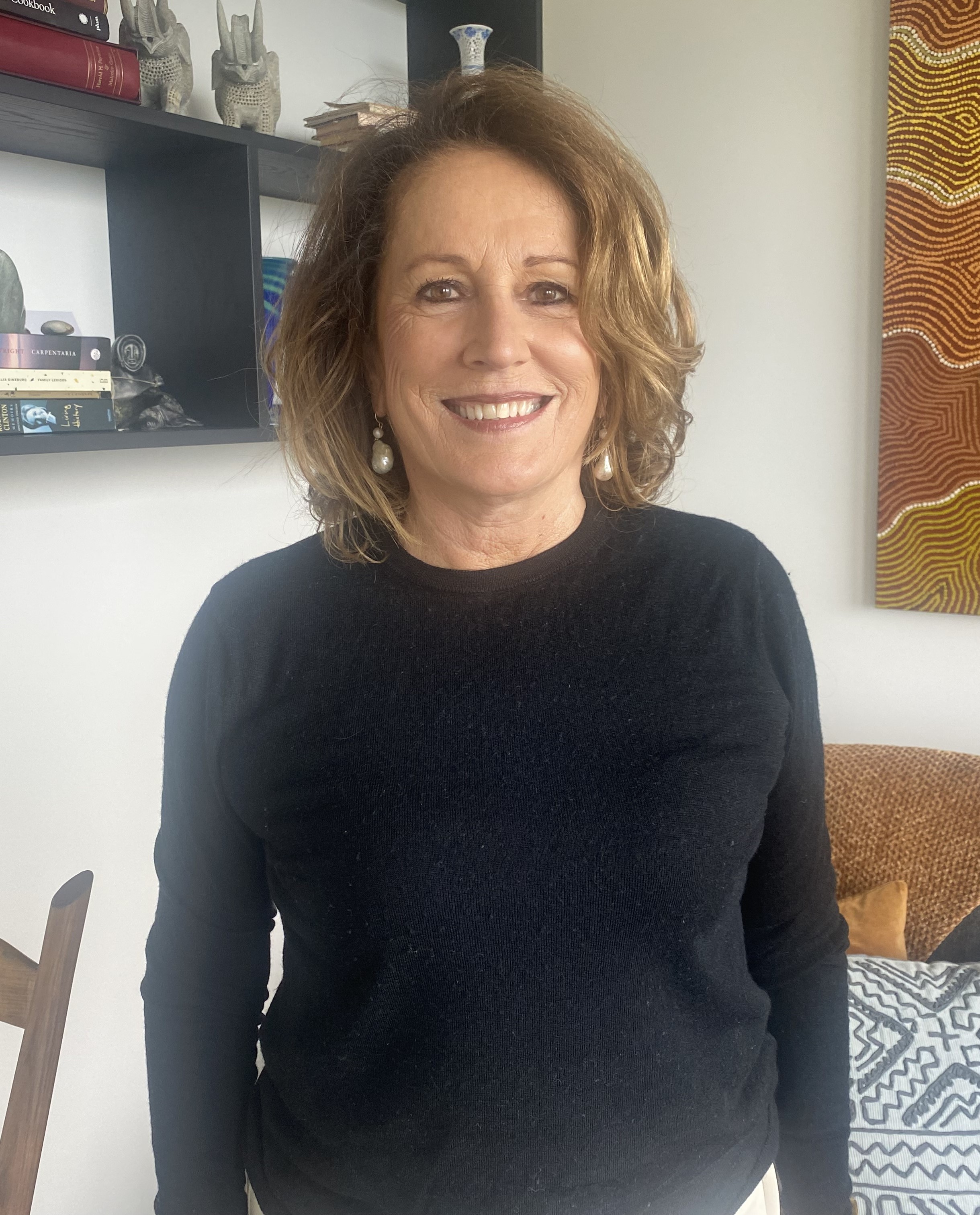 Woman in a black shirt is standing smiling at the camera. She is in a home setting which has a couch and indigenous artwork in the background.