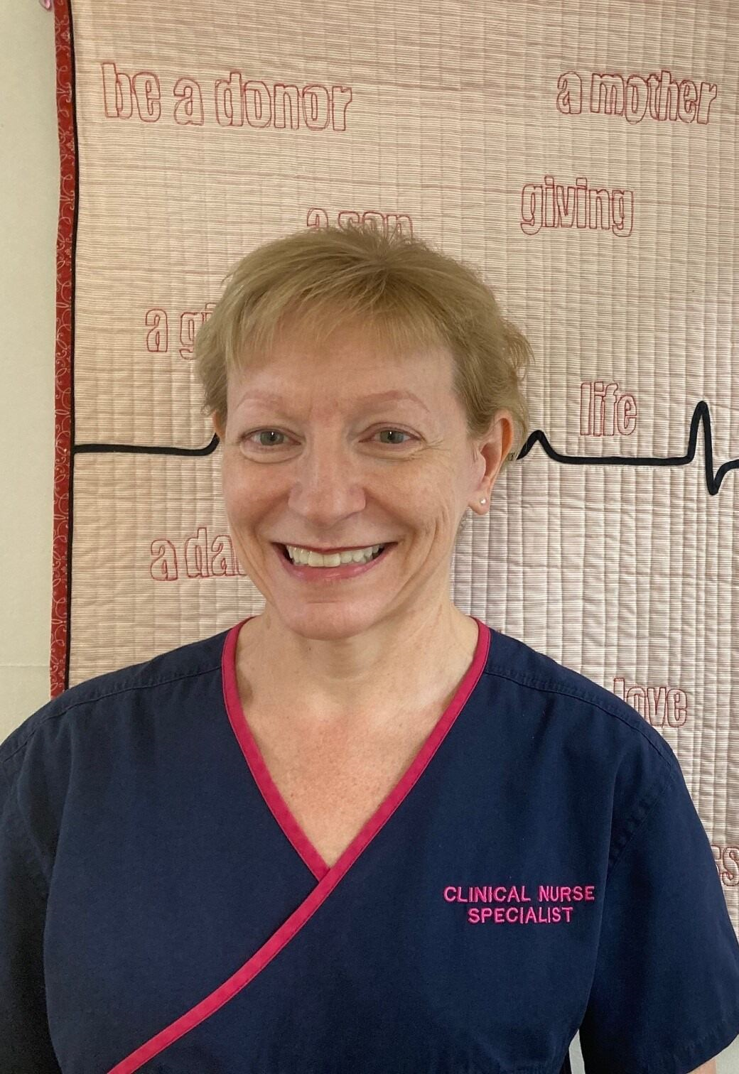 Woman in a nurse uniform standing in front of a hanging quilt. The quilt has words on it that refer to organ donation.