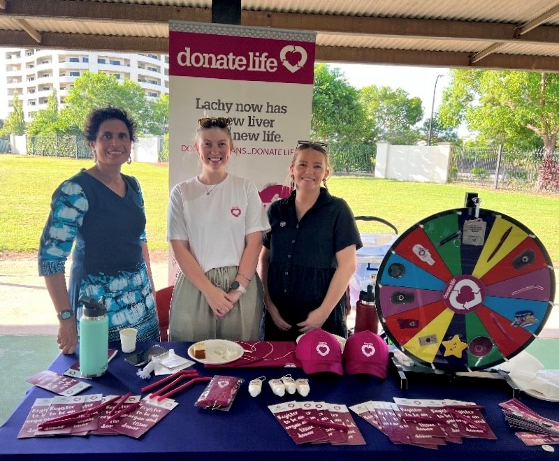 Three women stand behind a DonateLife stand at a festival.