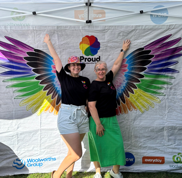 Two women pose in front of a pair of rainbow coloured wings at a Pride event.