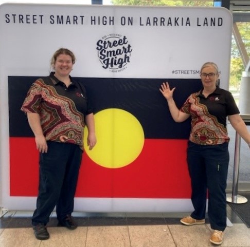 Two women in uniforms pose in front of the Aboriginal flag.