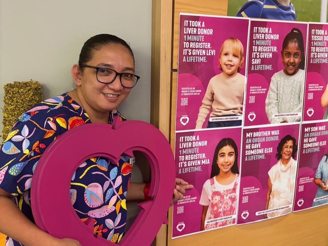 A female nurse holds up a wooden cut out of the DonateLife logo in front of DonateLife Week 2024 posters.