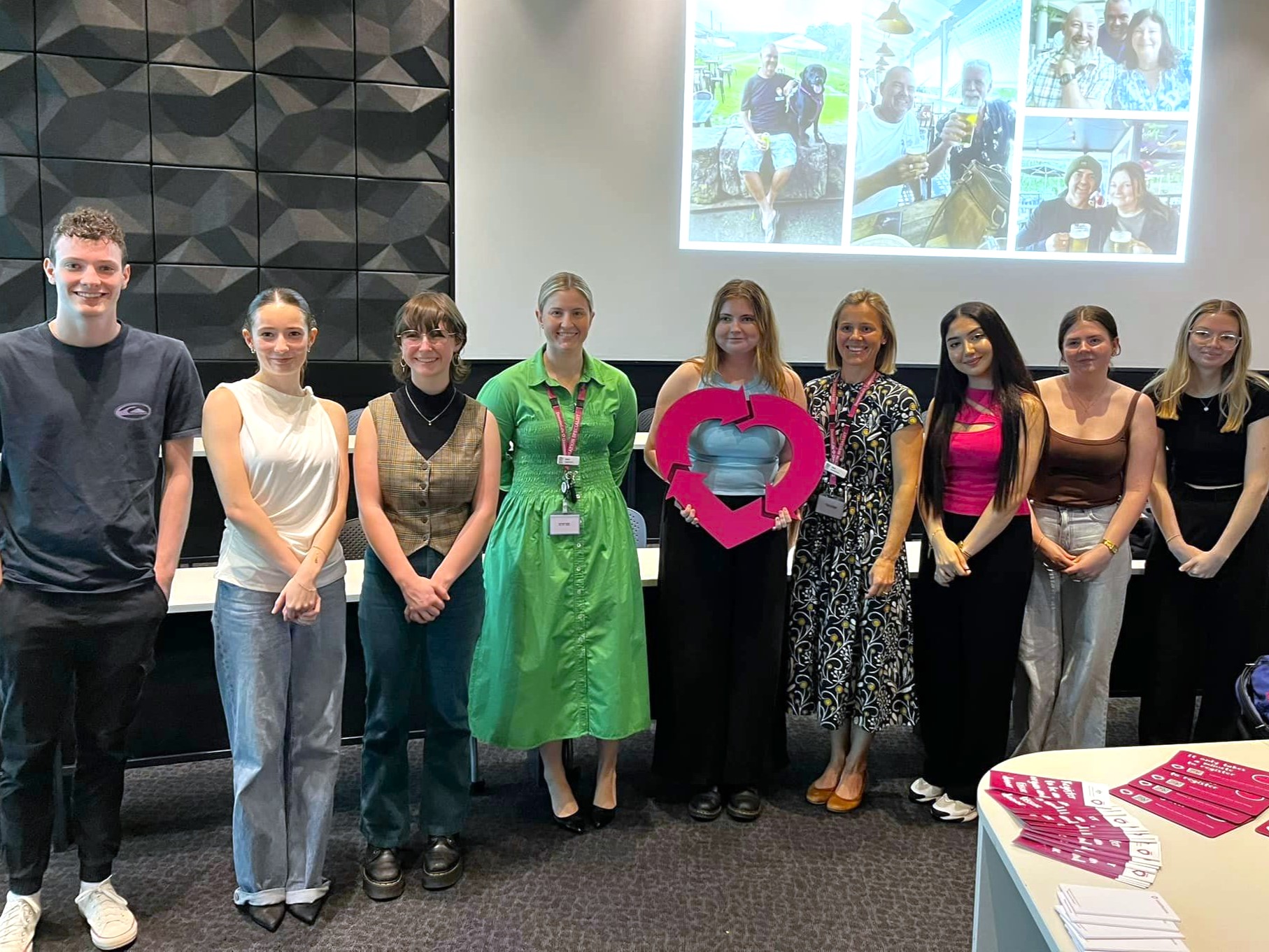 A group of university students pose at the front of a university lecture theatre. A young lady in the middle holds a wooden cut out of the DonateLife logo.