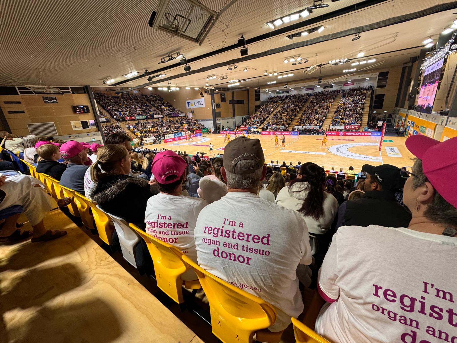 A view from the crowd in the stands of an indoor netball game being played.