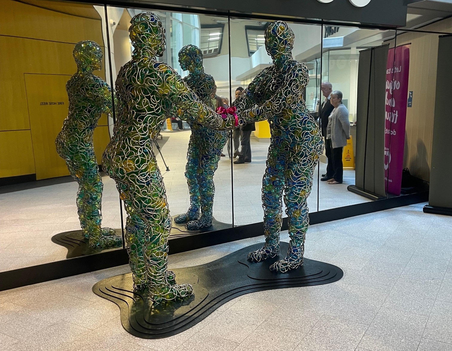 A glass and metal sculpture of two people standing facing each other and holding hands. It is in front of a mirror.