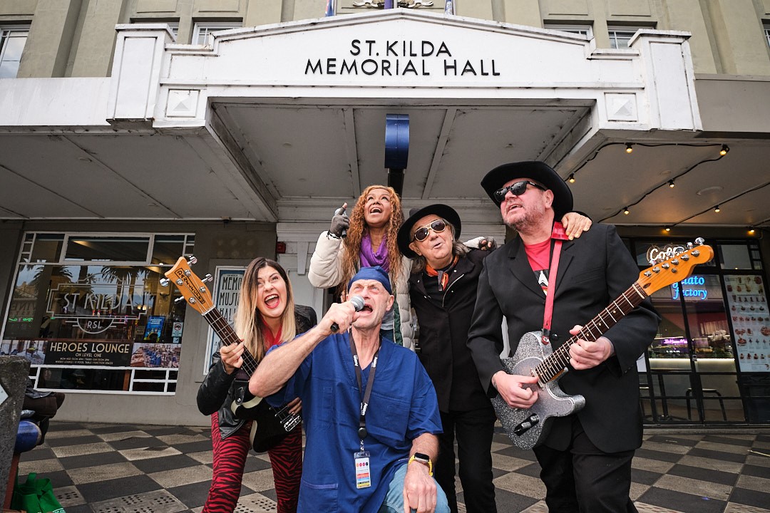 A band called The Oz Transplants pose with their instruments and a doctor in blue scrubs in front of St Kilda Memorial Hall in Victoria.