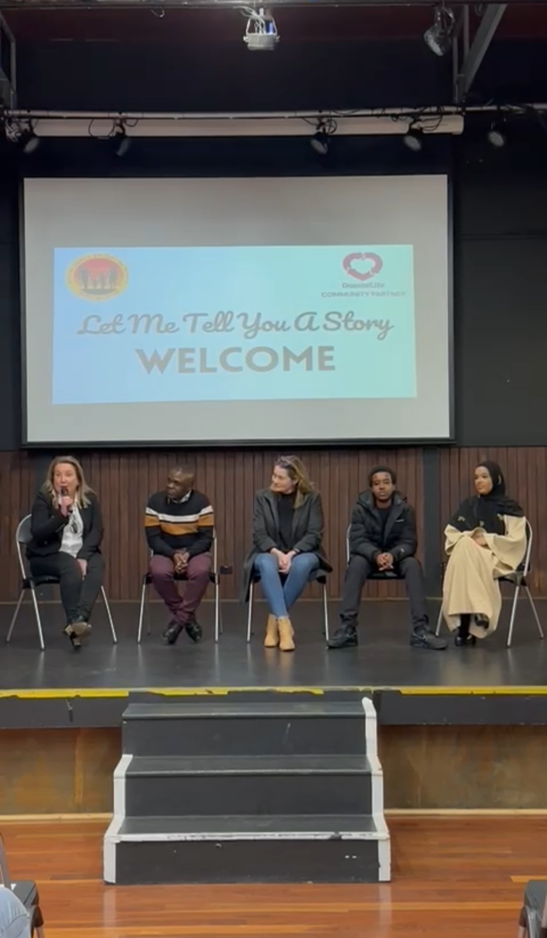 Five people sit on a stage, in the middle of a discussion. The woman on the left is speaking into a microphone and looking into the crowd, while the others are looking at her as she speaks.