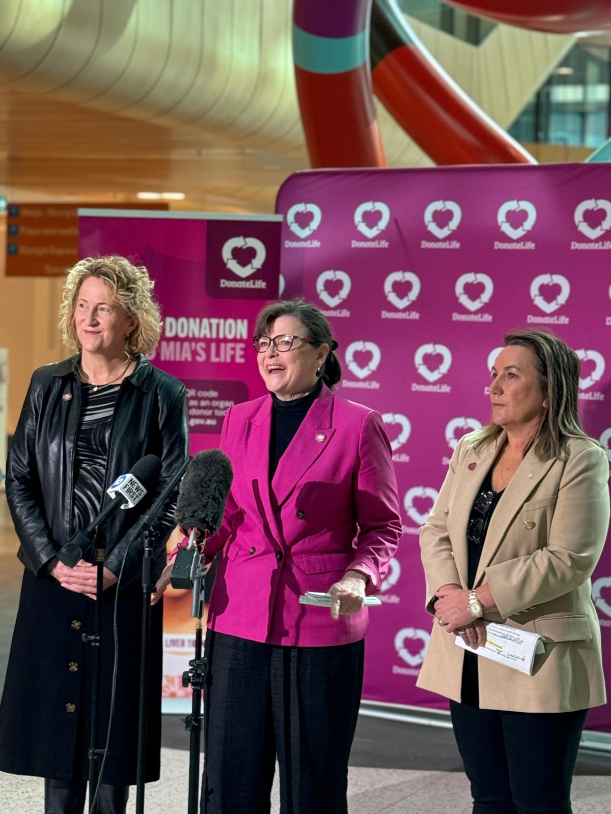 Three women stand in front of microphones, with DonateLife banners behind them. The woman in the middle is giving a speech. 