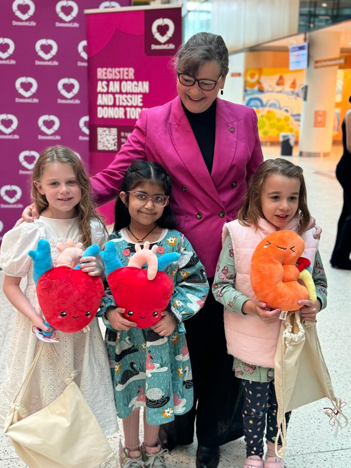 A woman stands with 3 children, holding organ plushies, as they smile for a photo together. The woman is looking down at the children and smiling. 