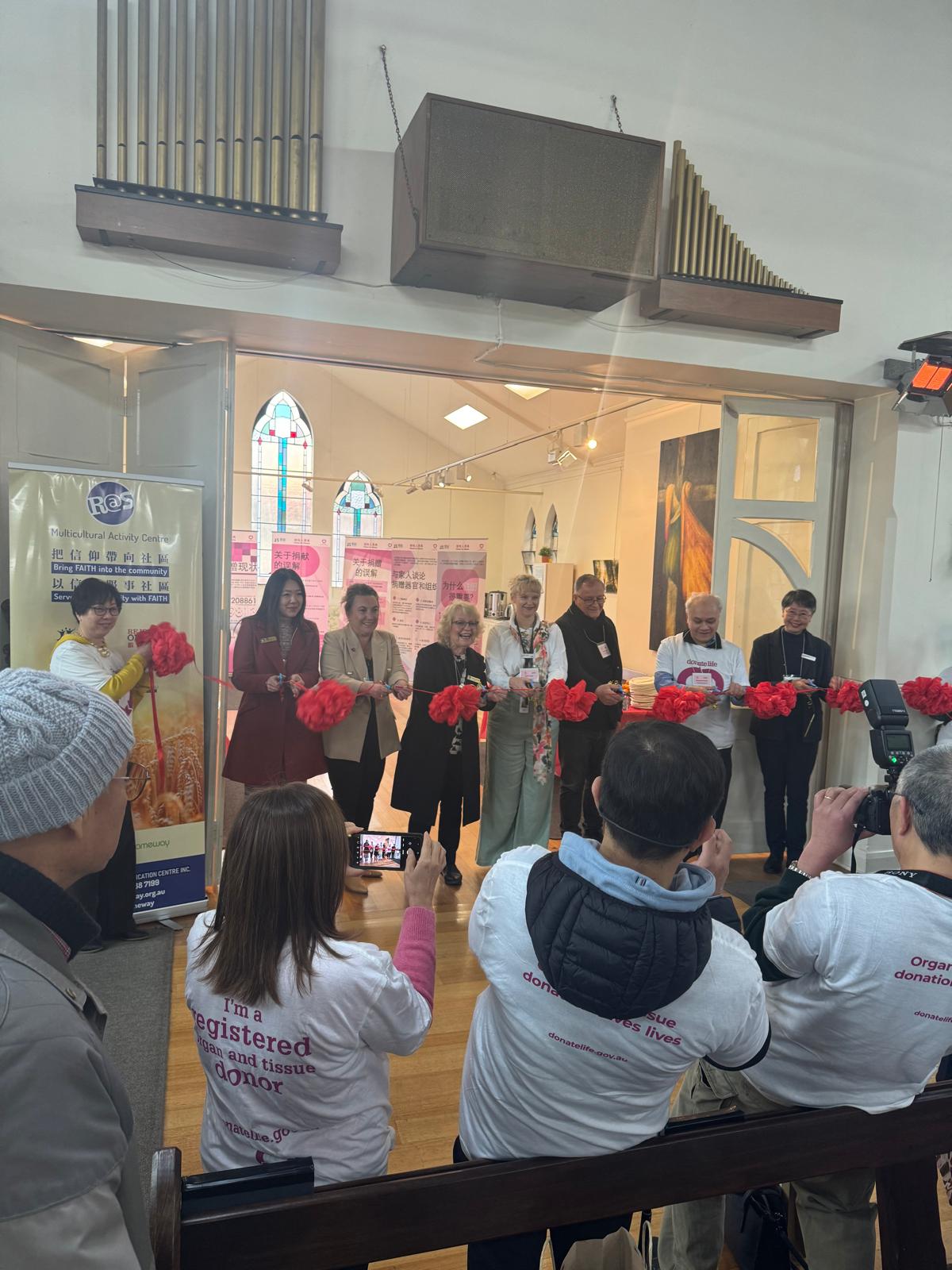 A group of people line up to cut a red ribbon strung across a large doorway. Others are grouped in front of them, taking photos of the moment.