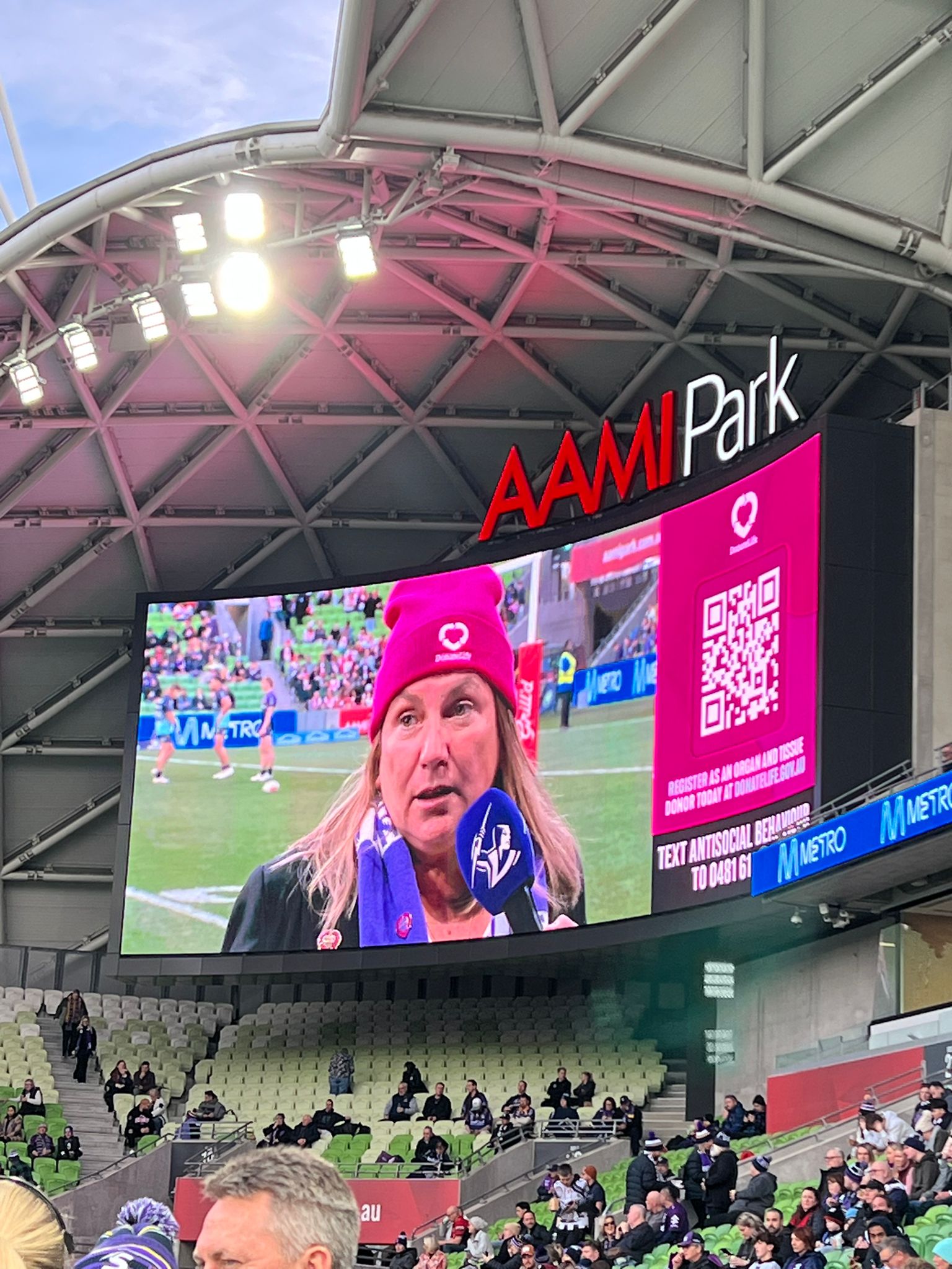  A photo from AAMI Park, which shows one of the big screens in the stadium. A woman in a magenta beanie is projected onto the screen. She is speaking into a blue microphone. 