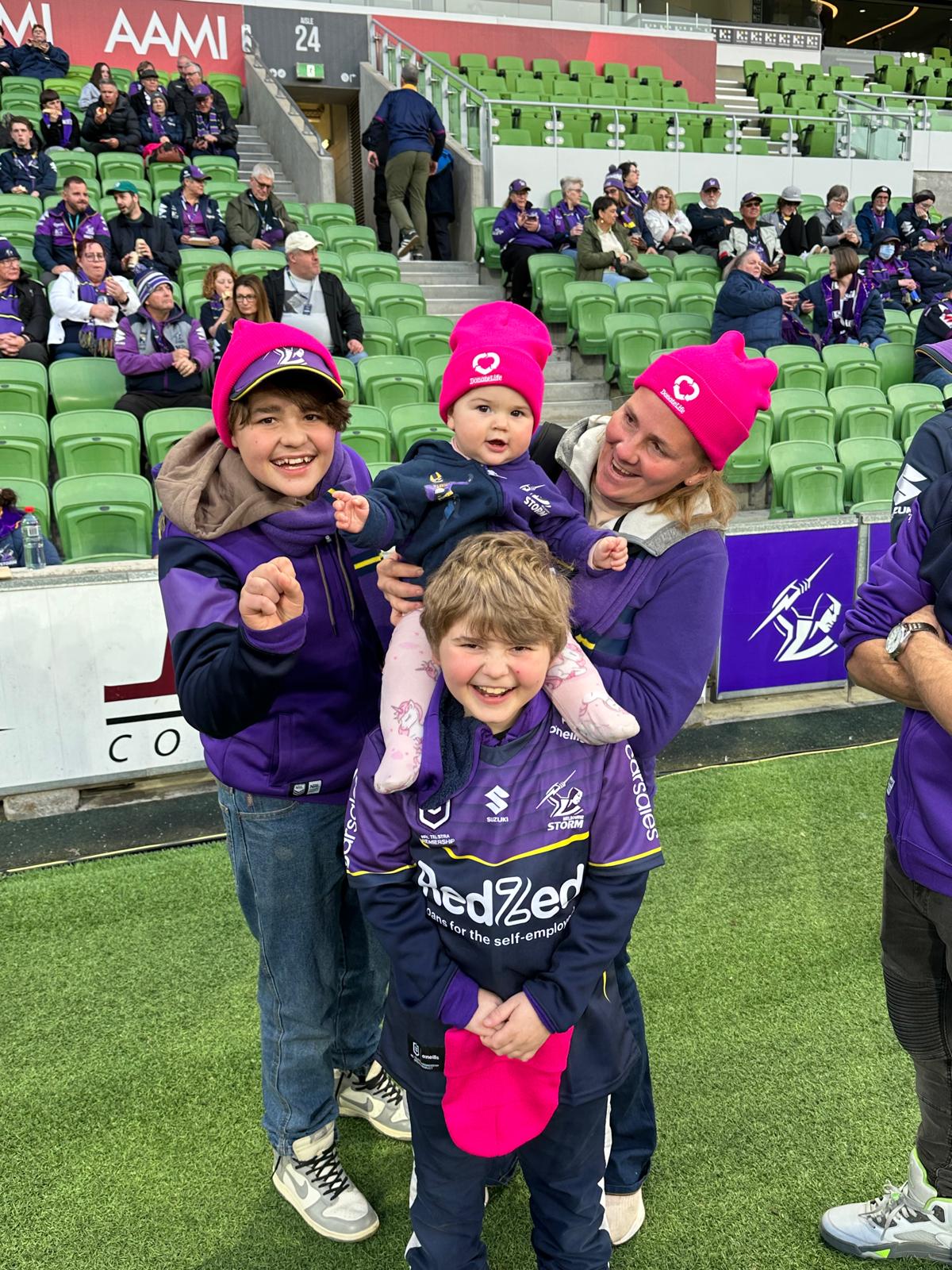 A family photo taken on the field at a sporting arena. Two young boys wear Melbourne Storm jerseys, a baby is sitting on one of the boy’s shoulders and wearing a magenta DonateLife beanie, and a woman also wearing a beanie holds onto the baby and is smiling at her. 