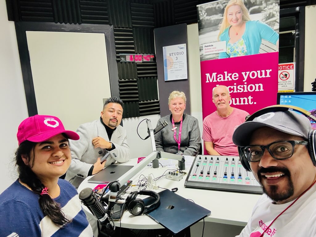 Five people sitting in a radio booth, sound board equipment and headphones on the table in the middle