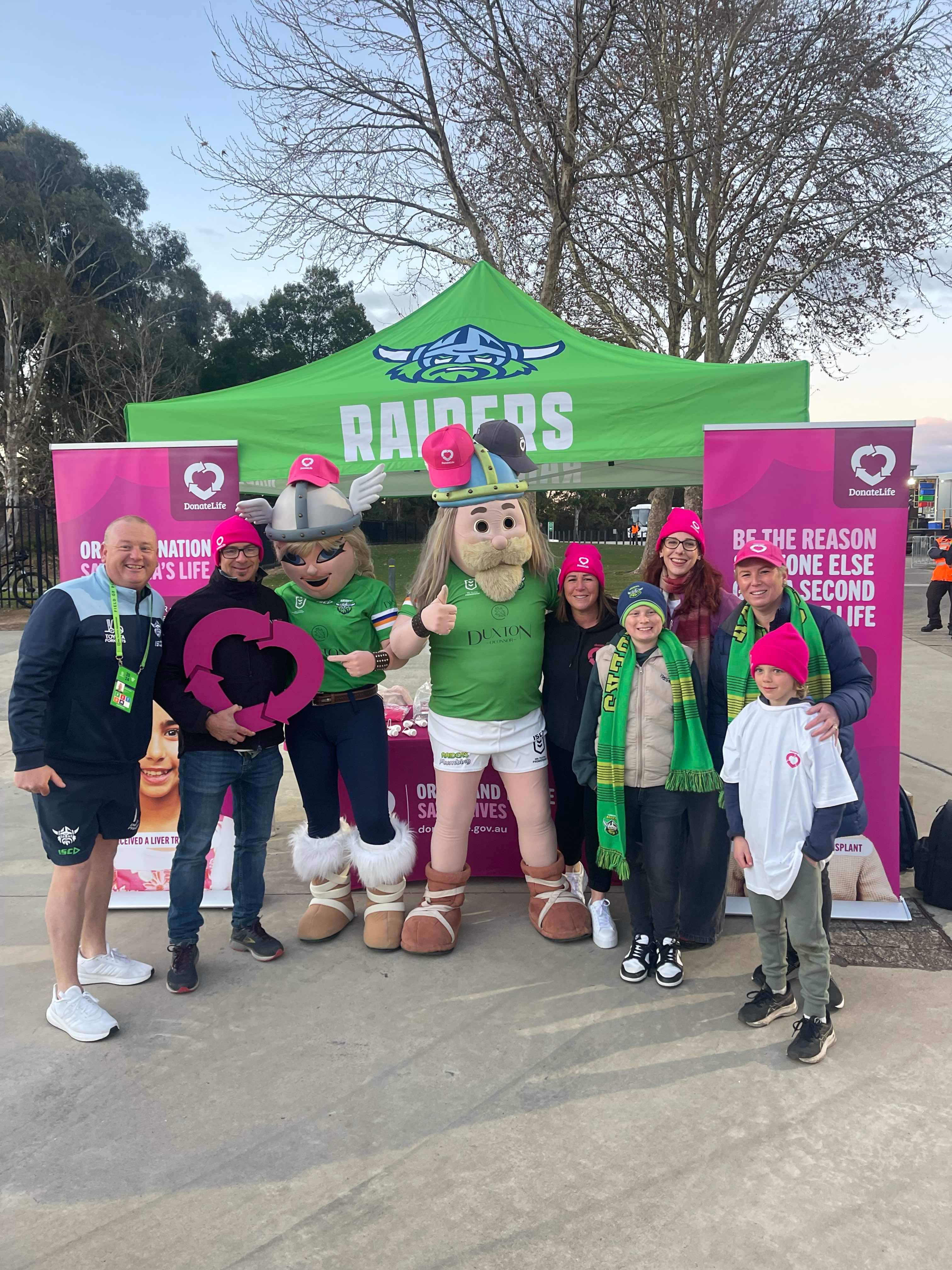 A group of people standing in front of a stall, which has the green Canberra Raiders logo on it and magenta DonateLife banners. Standing with the group are 2 people dressed as Canberra Raiders Viking mascots.