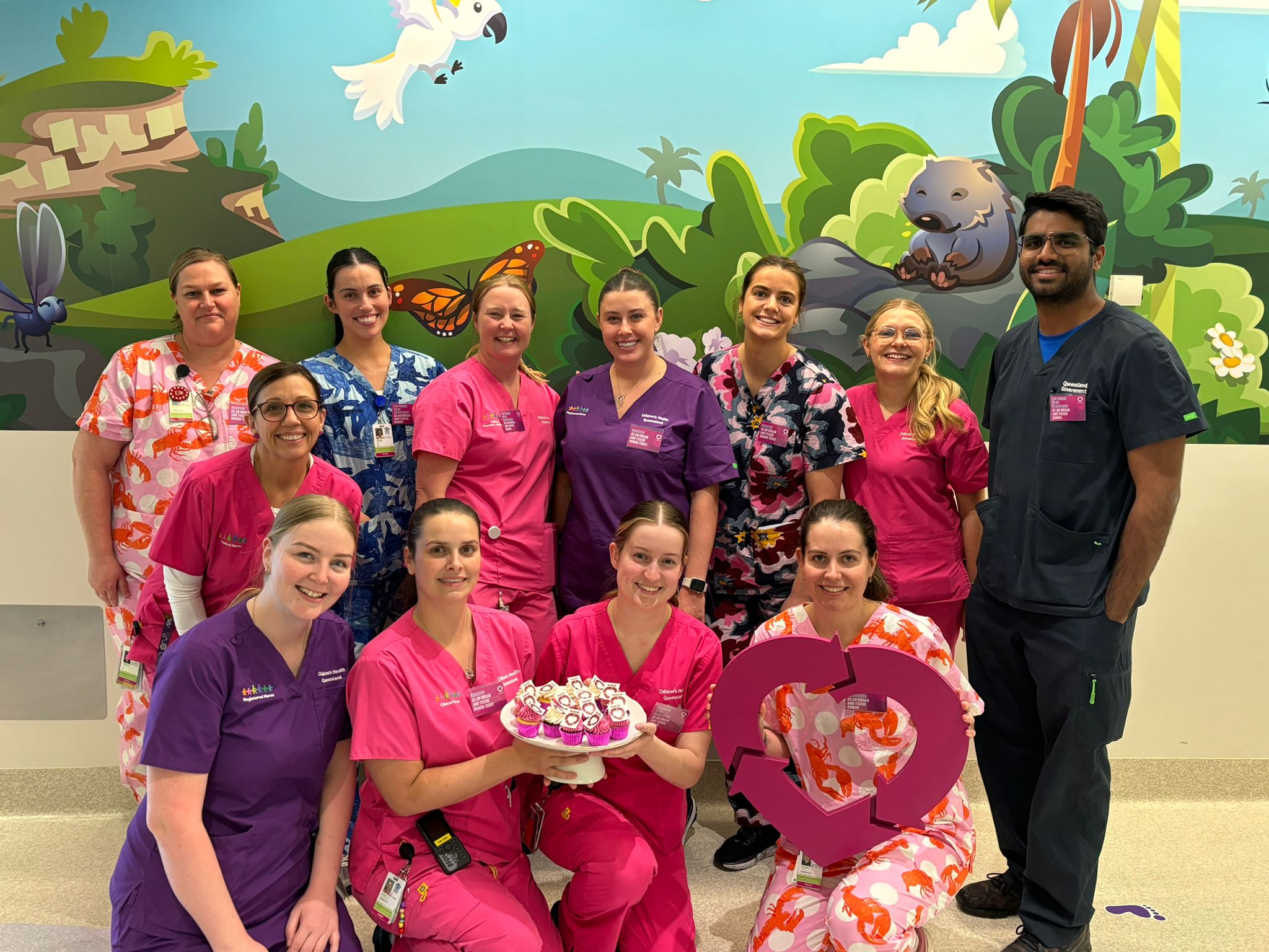 A group photo of nurses wearing a variety of pink and magenta scrubs, holding a plate of magenta decorated cupcakes and the DonateLife heart emblem.