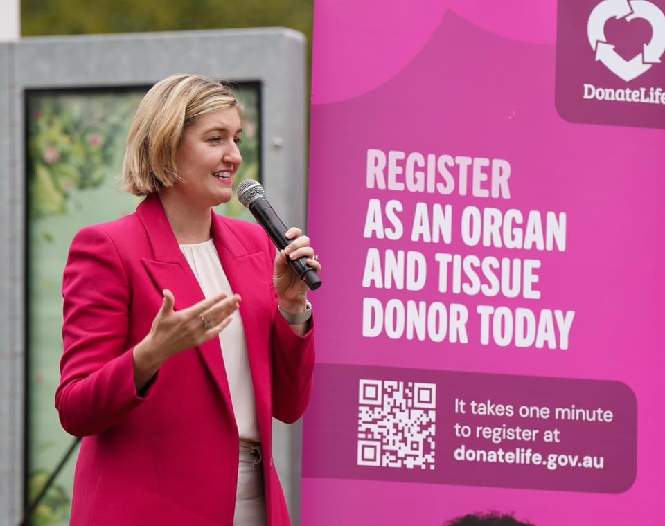 A blonde woman speaks into a microphone, a magenta DonateLife banner in the background.