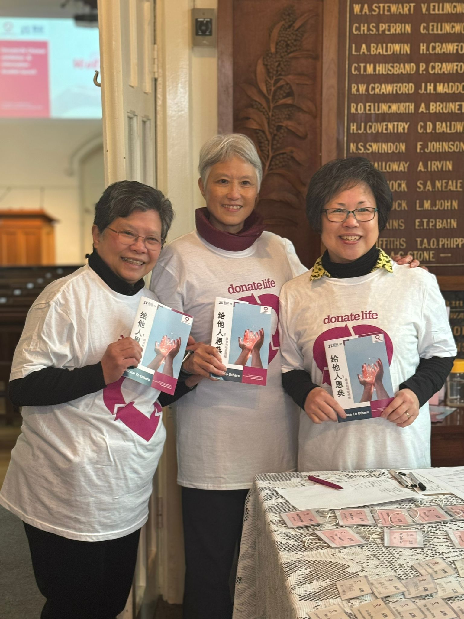 Three women of Asian background wearing DonateLife shirts hold booklets written in Chinese.