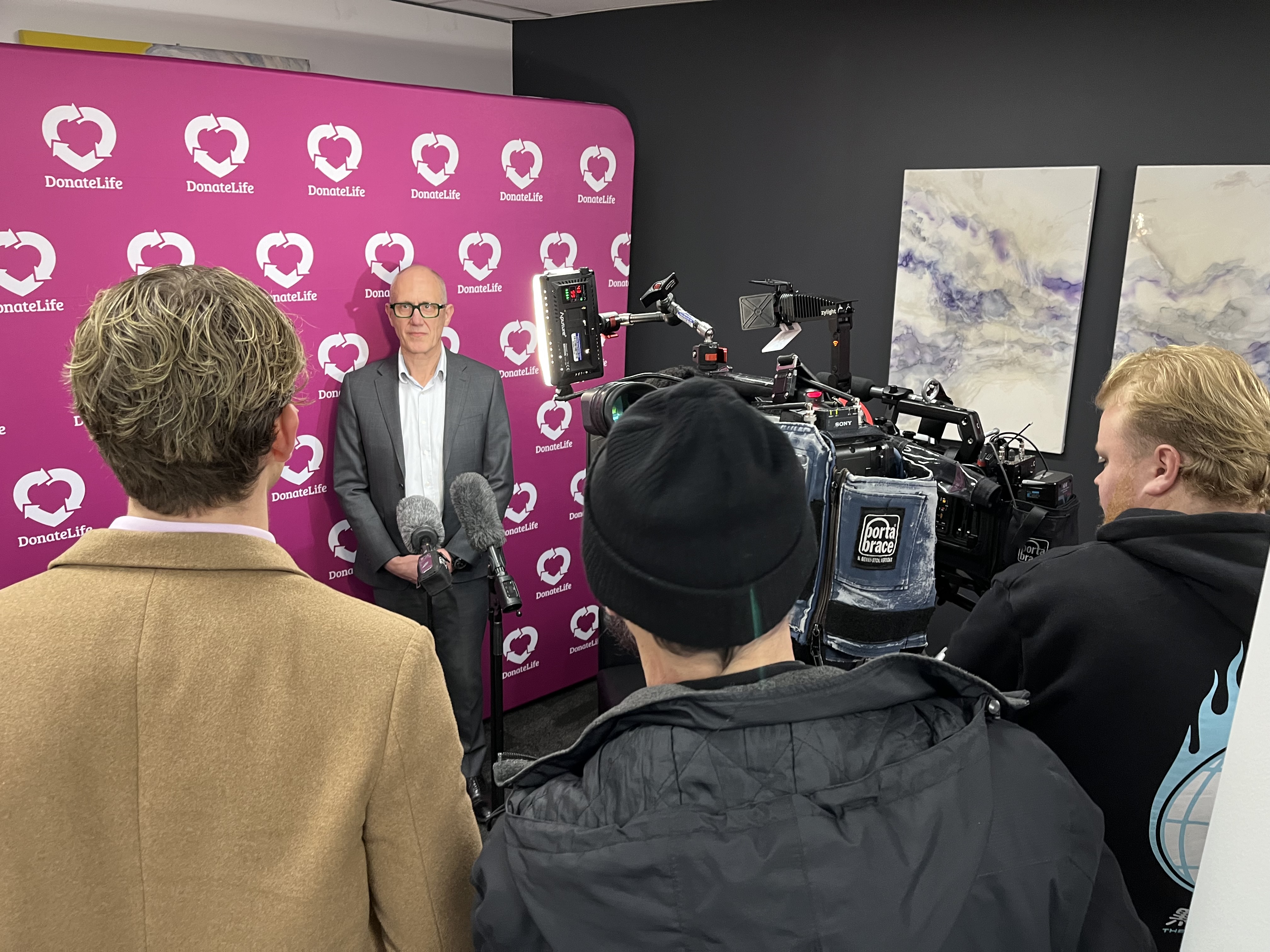 A man in a suit stands in front of a DonateLife branded banner, with reporters and a TV camera recording him as he speaks.