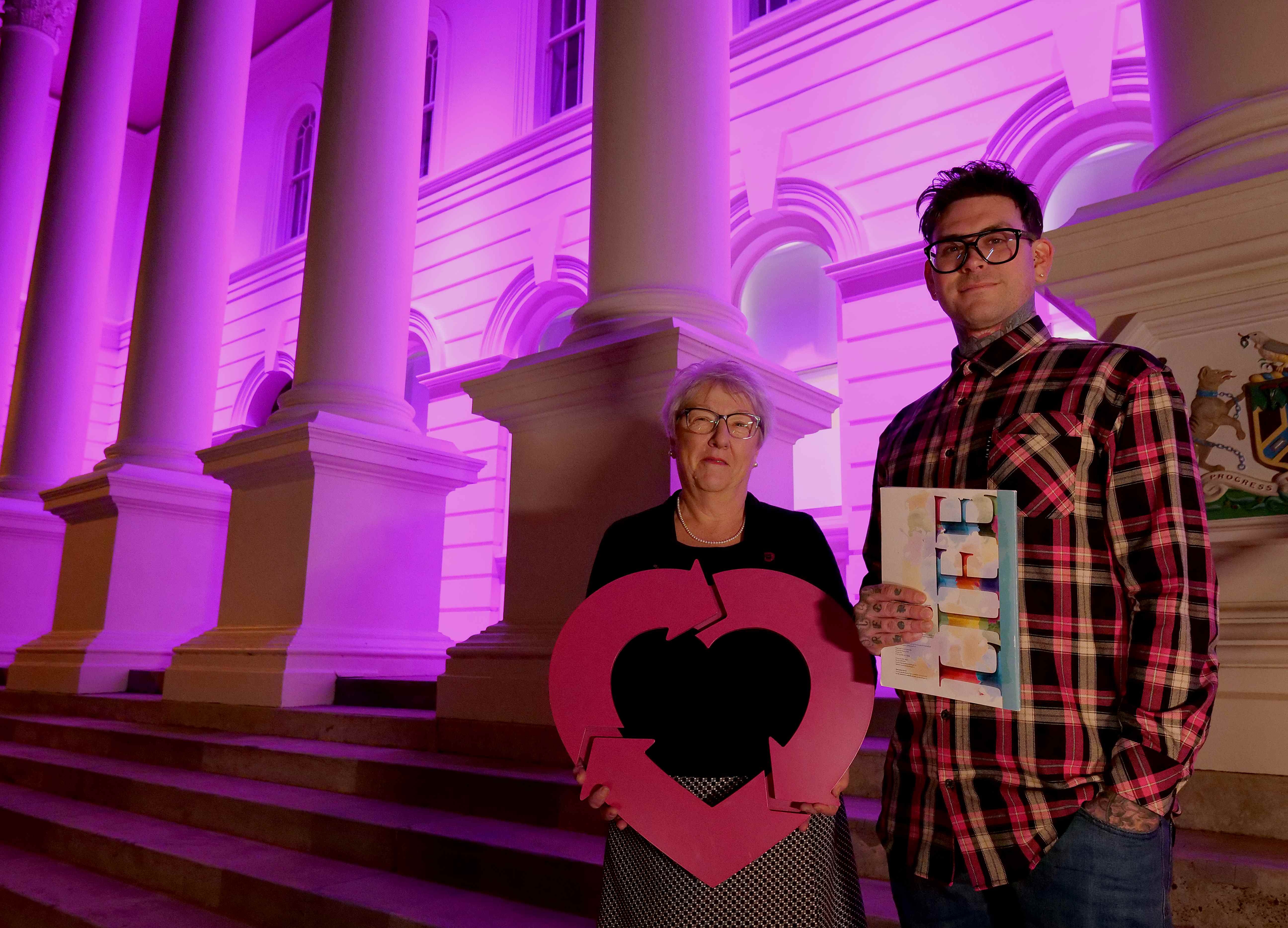 A woman holding the DonateLife heart emblem stands with a man in front of a government building lit up magenta.