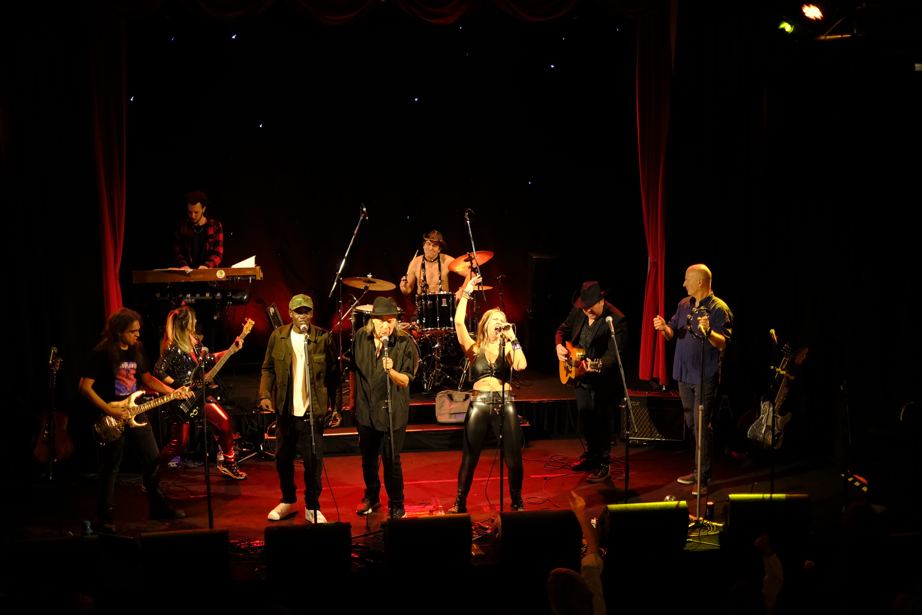 A woman in a black leather outfit sings on stage. She is joined by other singers and musicians playing electric guitars and drums.