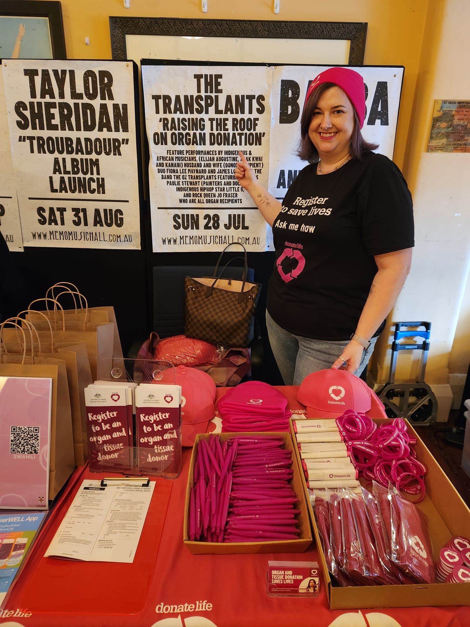 A woman in a magenta beanie and black DonateLife shirt stands behind a table with promotional merchandise.