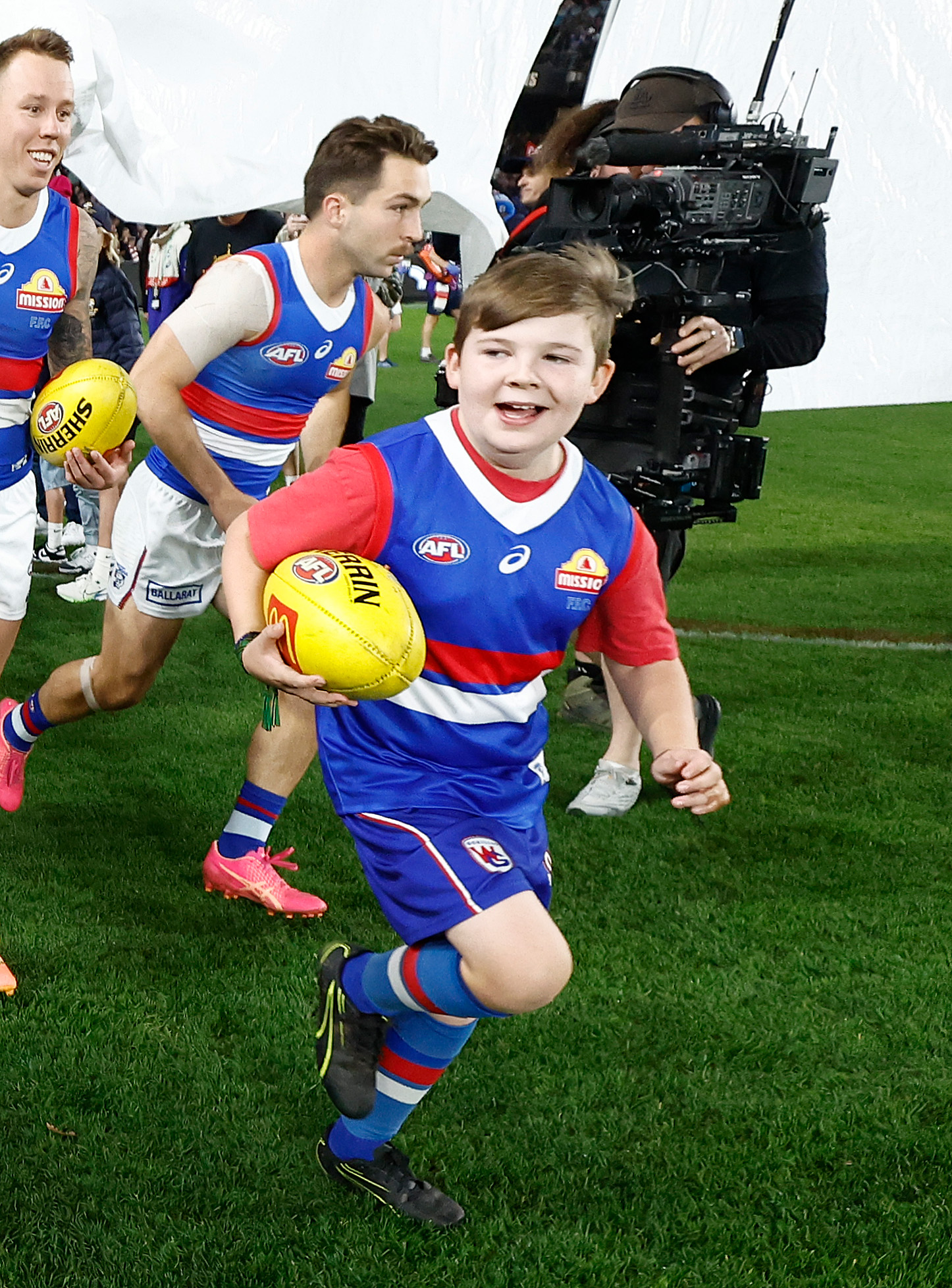 A young boy in Western Bulldogs uniform runs onto a field holding a football.