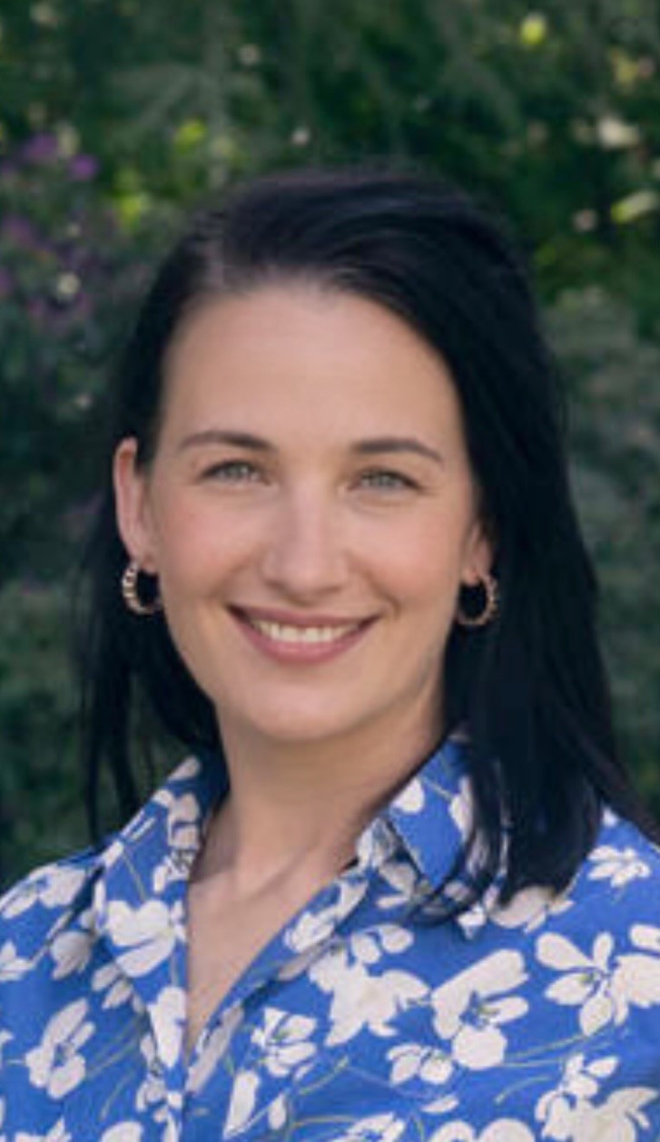 Woman with black shoulder length hair, in a blue floral top, smiling at the camera