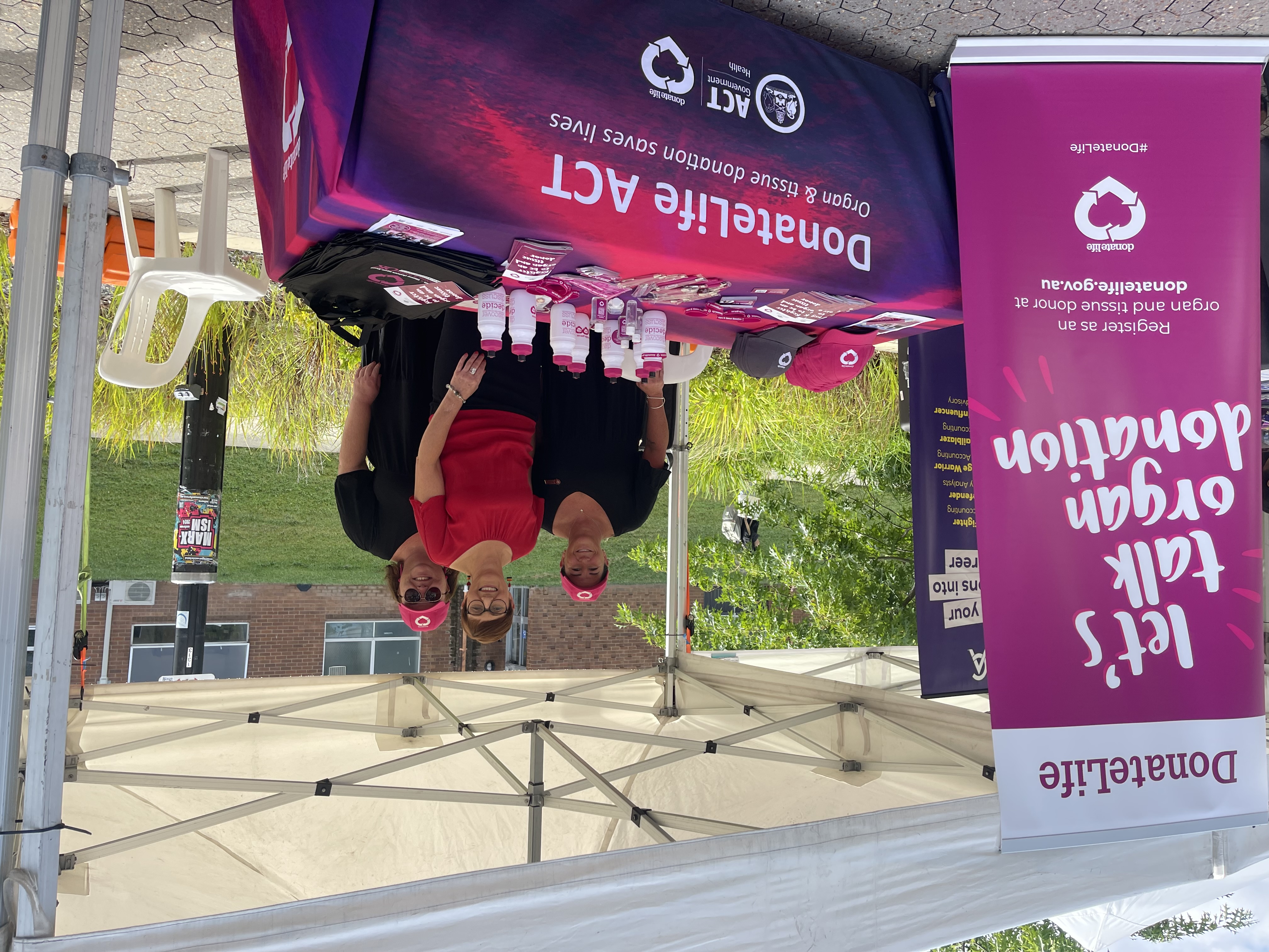 Three people standing at a marquee stall with DonateLife ACT branding, with a standing banner to the side that says ‘DonateLife: Let’s talk organ donation’. 