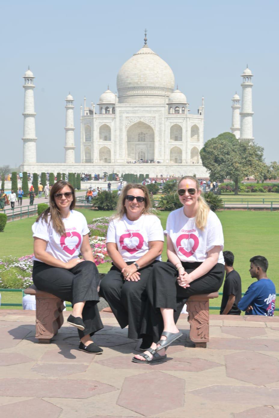 Three women in DonateLife branded shirts pose sitting on a bench with the Taj Mahal in the background.