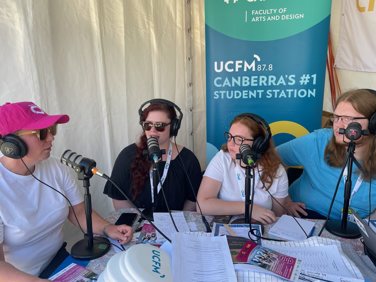 Four people sit at a table in front of microphones. They are in the middle of a radio interview.