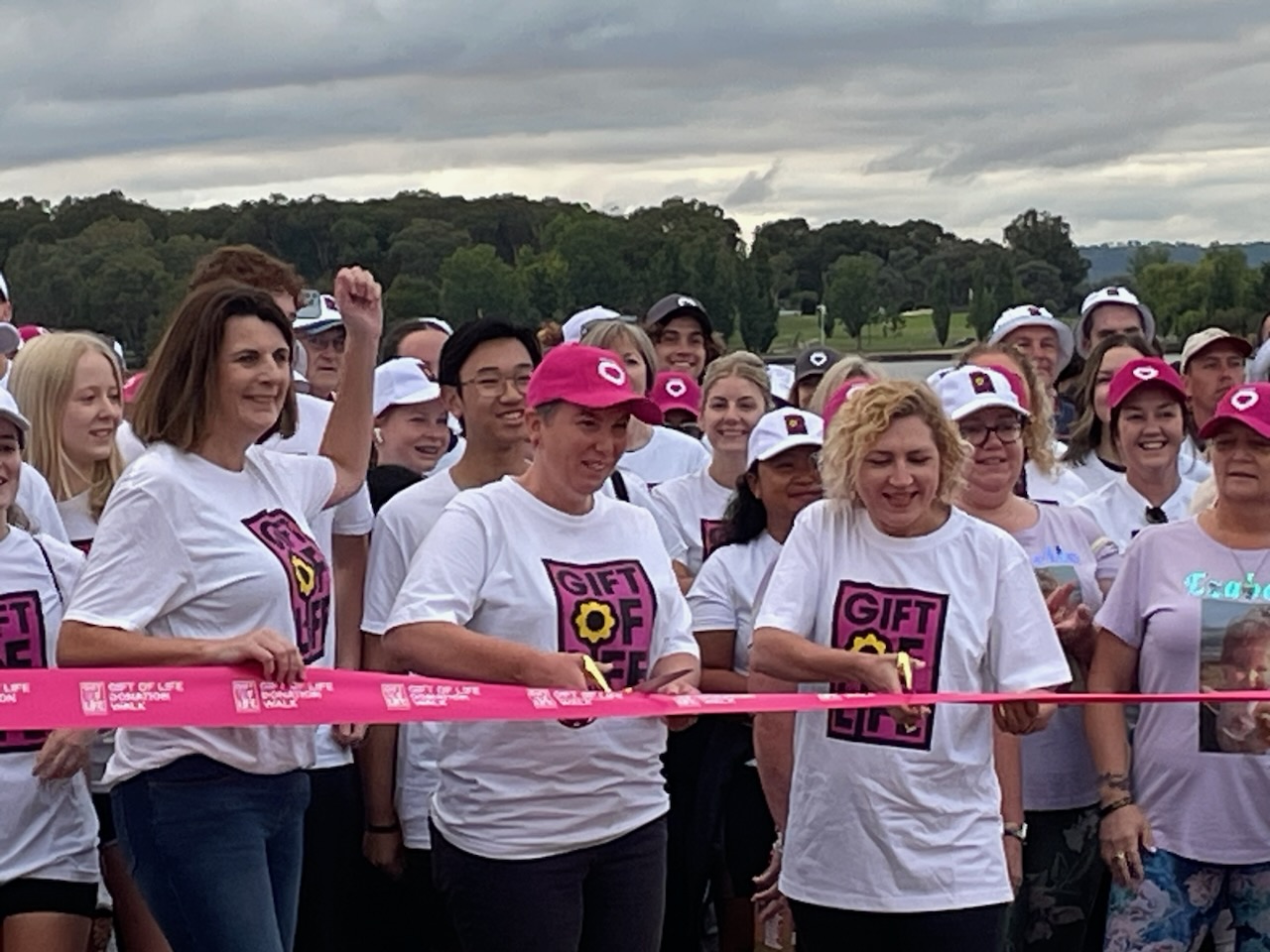 Participants at the Gift of Life Walk gather at the walk’s starting ribbon. The group are wearing Gift of Life branded shirts and DonateLife caps, and two women are cutting the starting ribbon with scissors