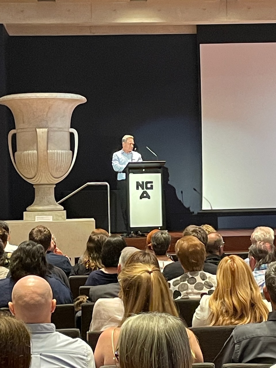 A man stands at a podium on a stage, delivering a speech to crowd, some of whom we can see sitting in the audience in front of him.