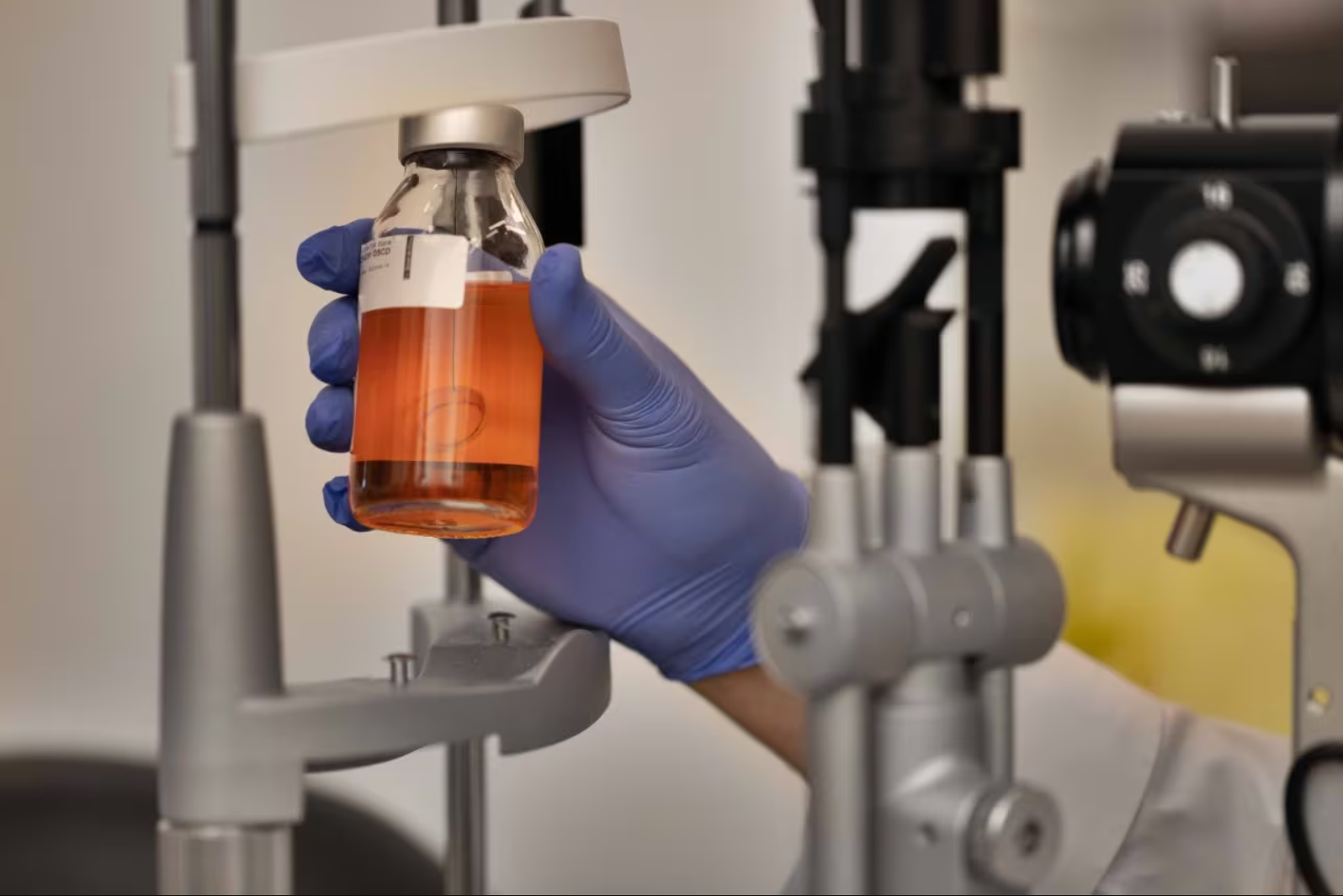 A hand in a blue glove holds a glass specimen jar containing an artificial cornea up as if inspecting it.