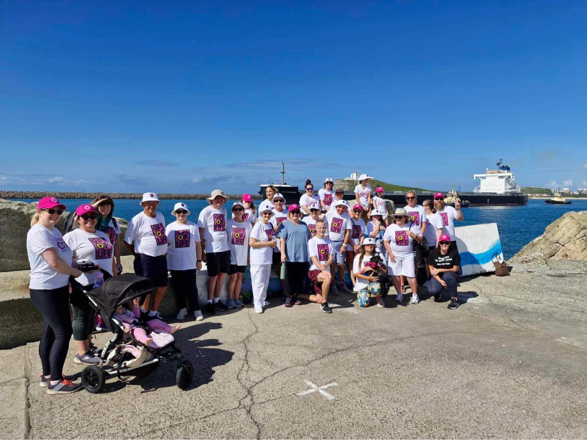 A group photo of participants at the Gift of Life Walk. They are posing on a waterfront, with the ocean and some boats behind them. They are wearing branded Gift of Life shirts and DonateLife hats.