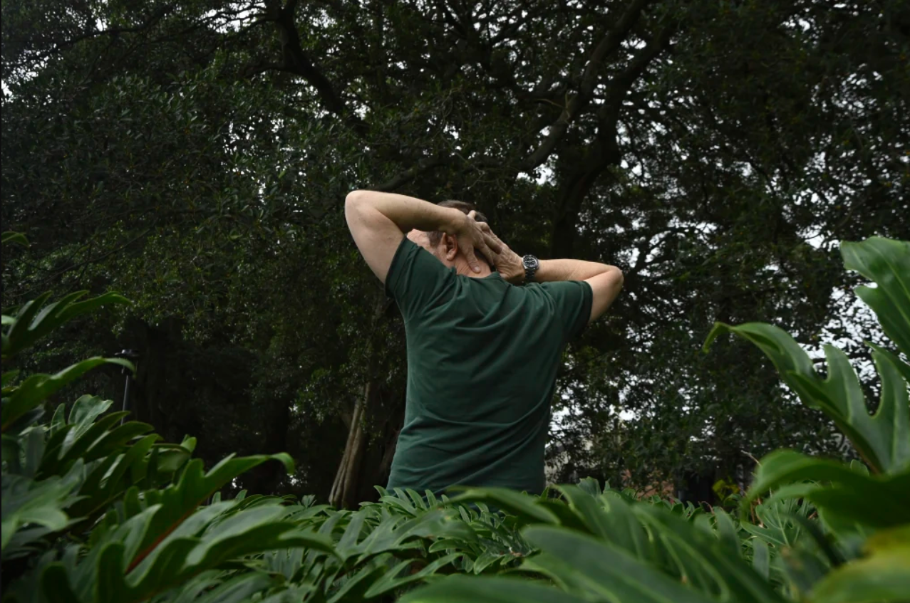 A man faces away from the camera so we only see his back. He is standing in a park with trees and plants framing him, and he stretches with his arms behind his head. 