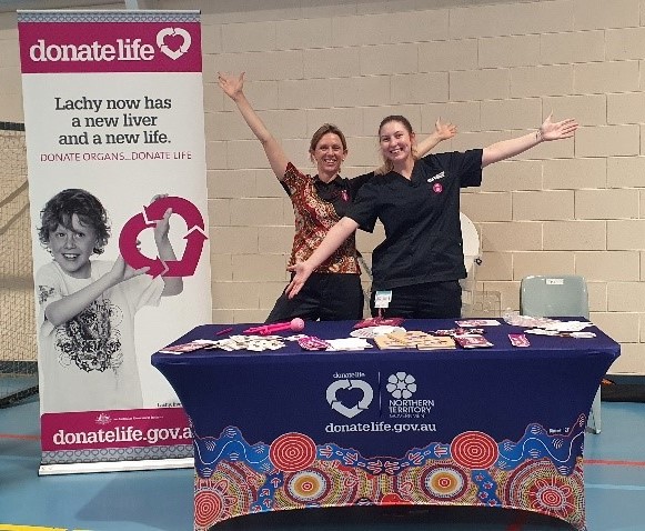 Two women stand behind a DonateLife NT stall with a DonateLife standing banner next to them. Their arms are thrown wide and they are smiling broadly.