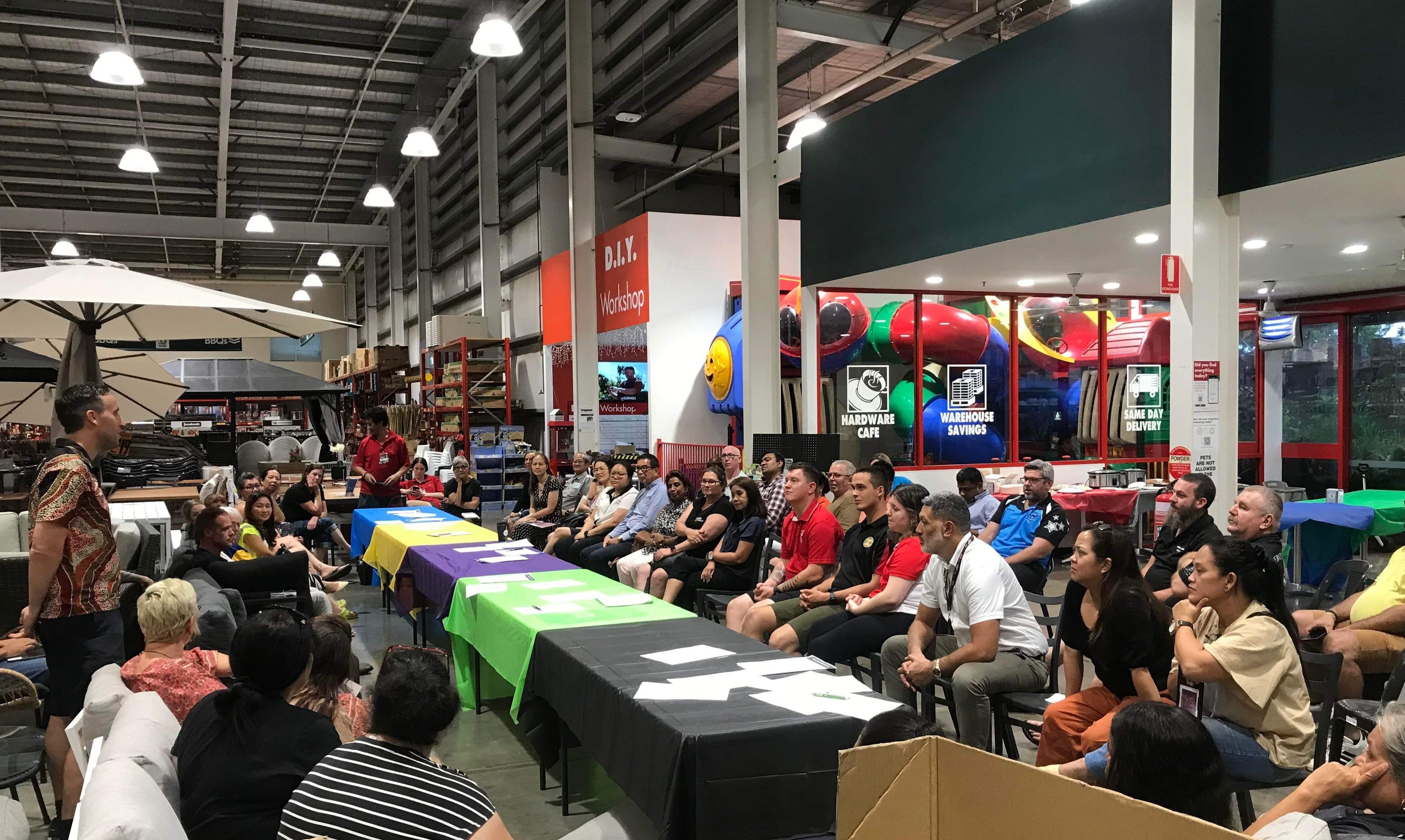 A group of people seated inside a Bunnings store, watching and listening to a man who is standing and speaking.