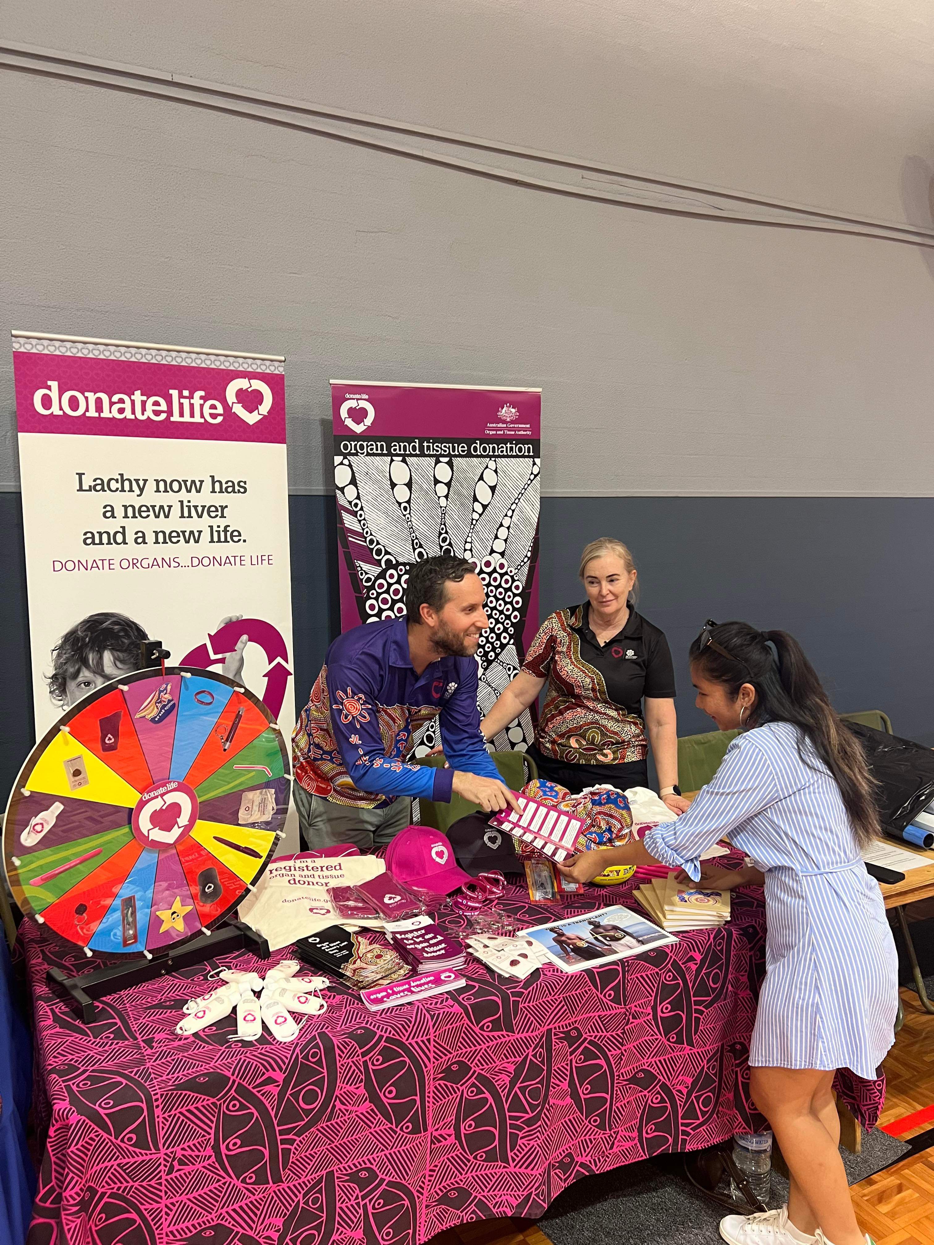 Two DonateLife staff members at a stall with DonateLife banners, merchandise and information, engaging with an attendee across the table.