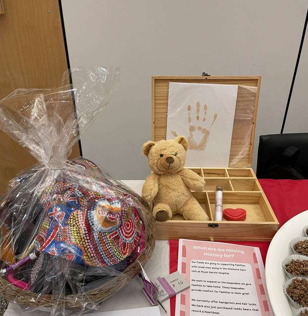 Various items that are being considered for inclusion in a bereavement pack laid out on a table – there is a teddy bear, a wooden compartment box, a wrapped-basket of DonateLife branded products, and a raffle sign-up with tickets.