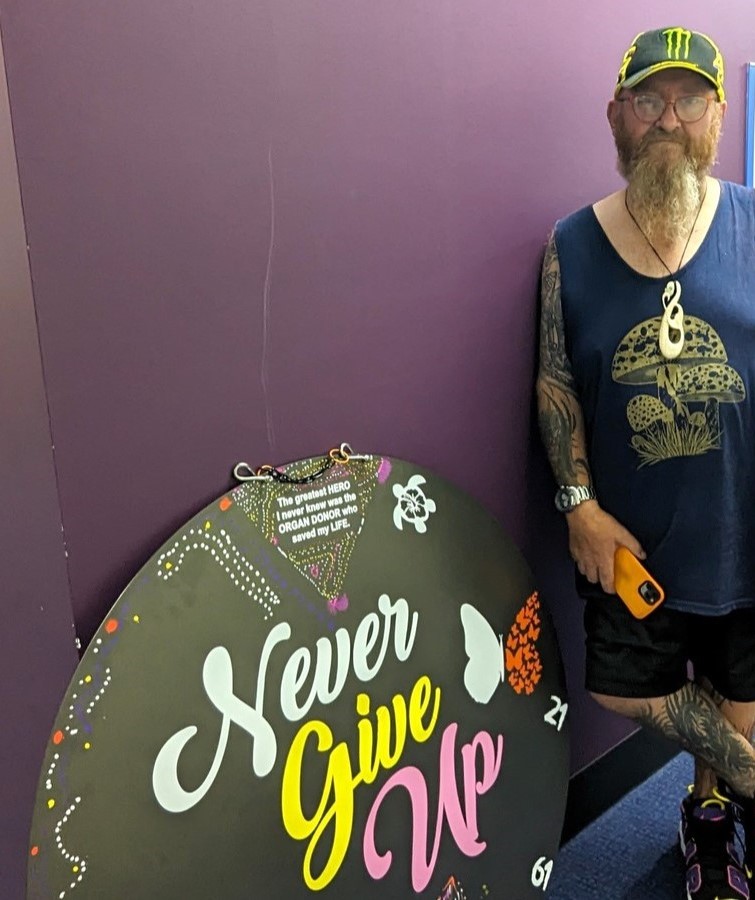 A man stands next to artwork he has created. It is a large, round piece of art that has the phrase ‘Never Give Up’ in the centre in white, yellow and pink cursive writing. The artwork features pictures around the sides, including a turtle with a flower on its back and two different butterflies.