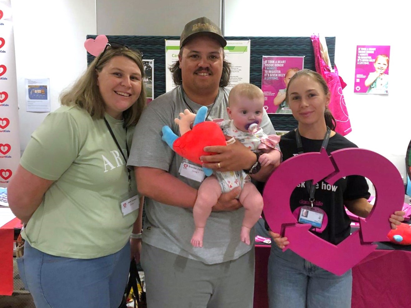 Three people stand together for a photo, the middle man holding a young baby. The woman on the right holds a magenta DonateLife heart emblem.