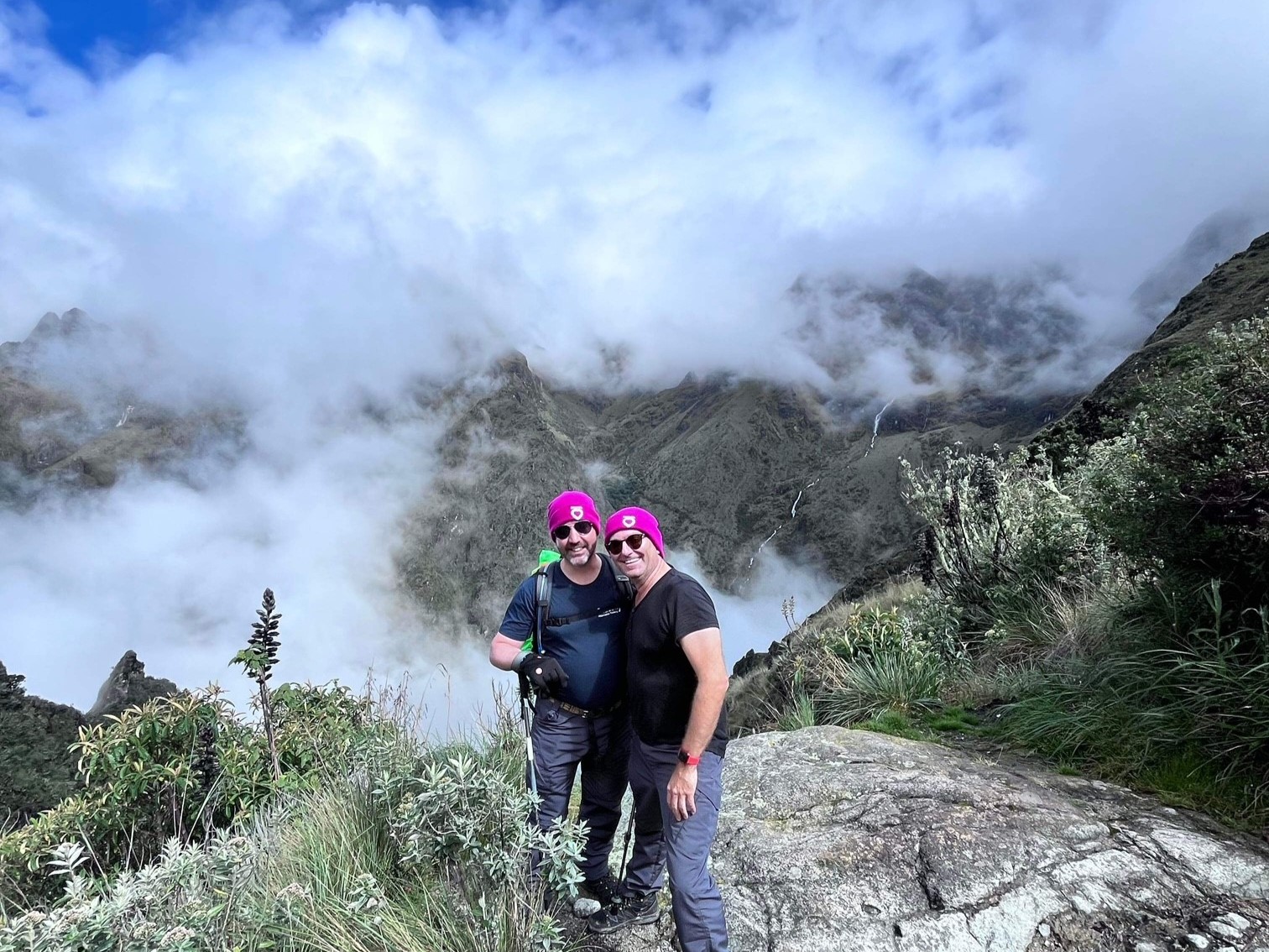 Two men in the middle of a hike, wearing magenta DonateLife beanies, pose on a mountain together, clouds and greenery in the background behind them.