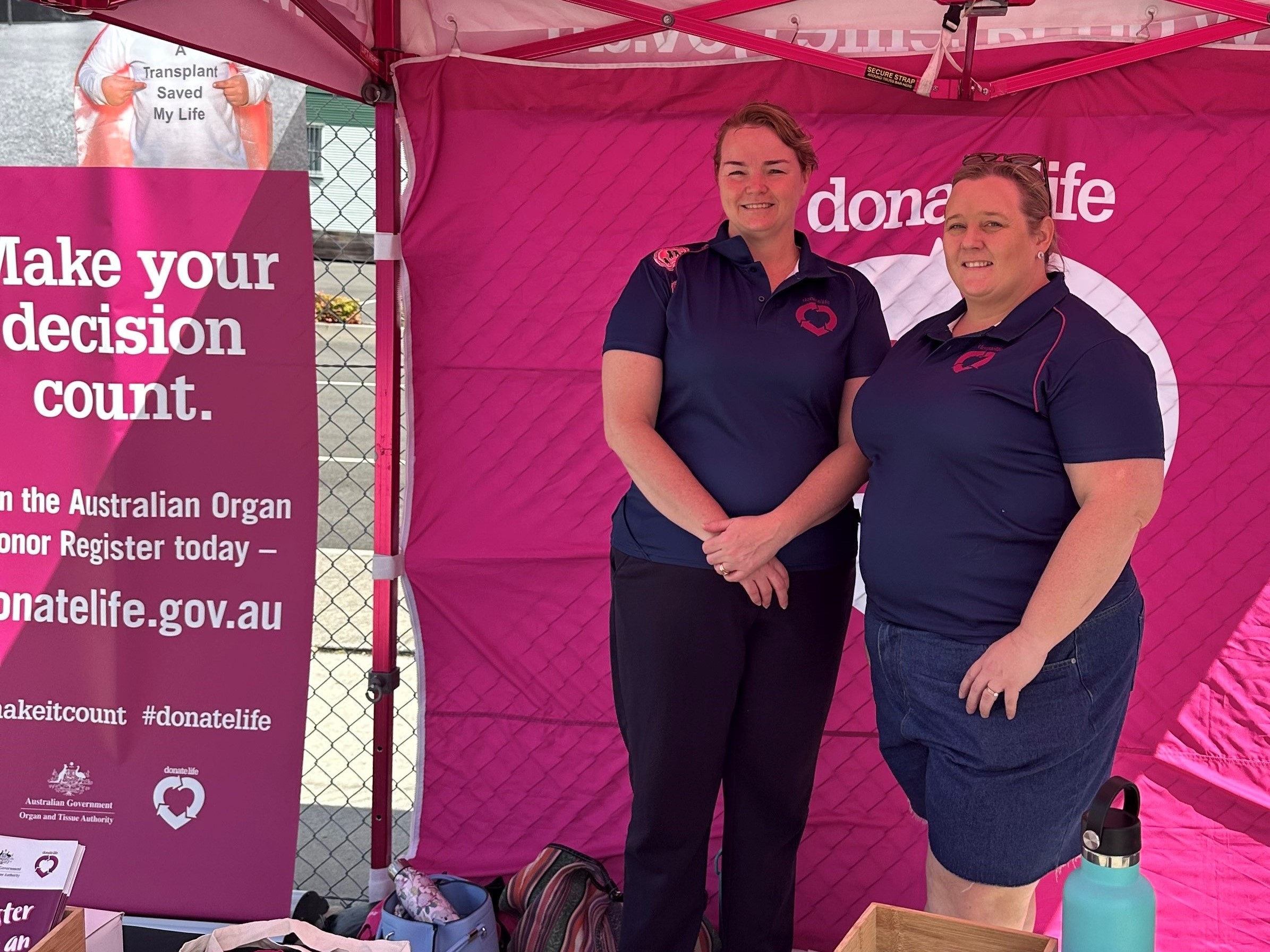 Two women in DonateLife t-shirts stand inside a DonateLife stall, smiling at the camera. 