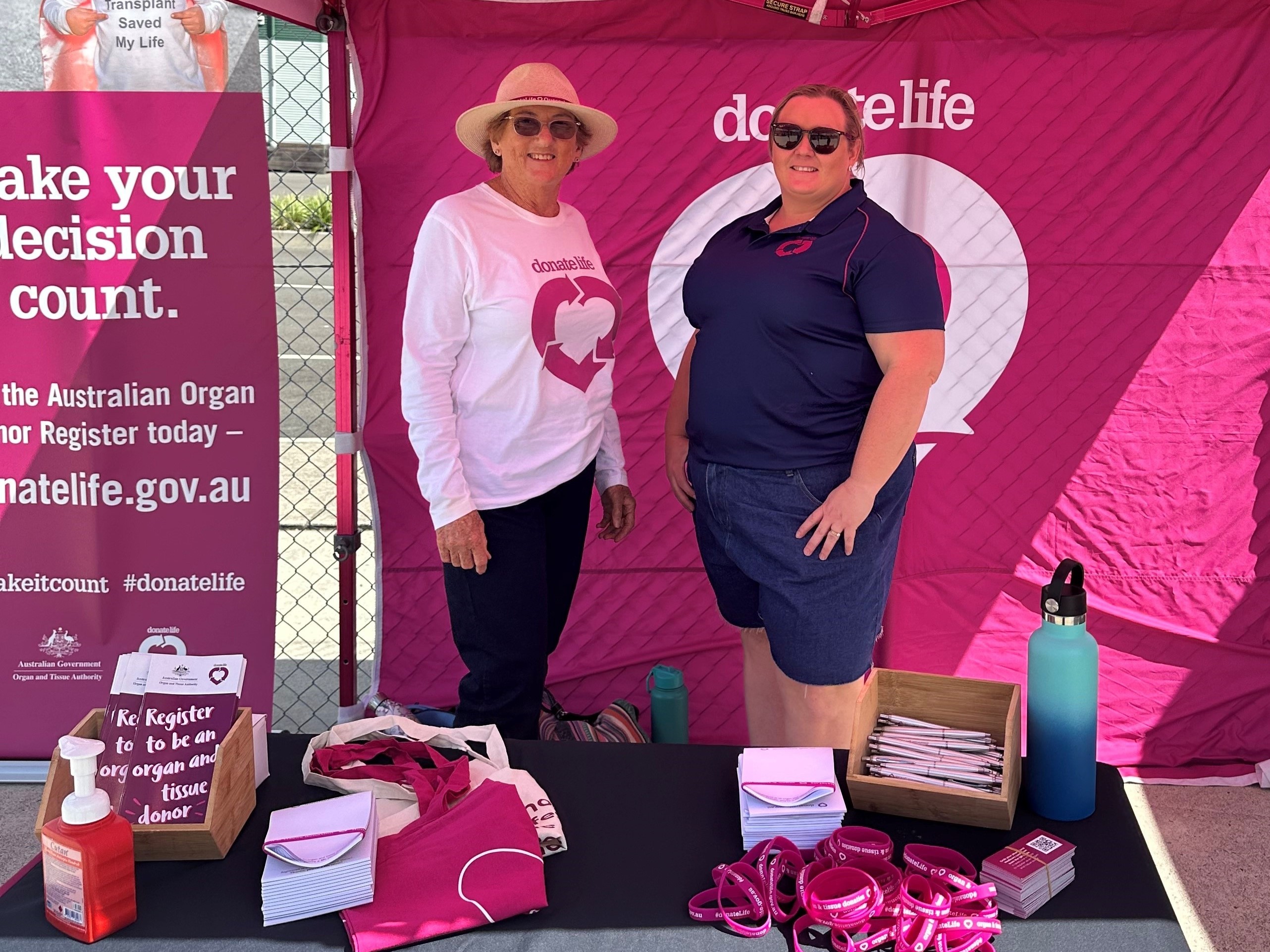 Two women in DonateLife t-shirts stand inside a DonateLife stall, smiling at the camera.