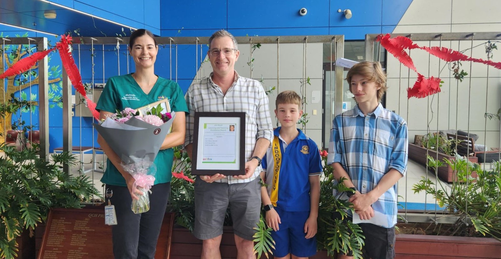 Two adults and two children pose for the camera, holding a bouquet of flowers and a framed award.